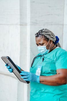 Focused nurse in scrubs using a tablet, wearing a mask for safety.
