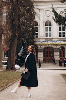 Smiling woman in graduation gown holding diploma in front of a historic building.