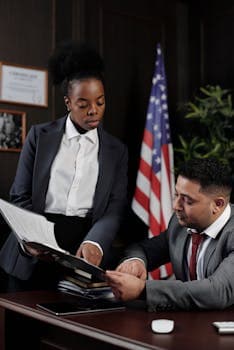 Two professionals reviewing documents in an office with an American flag in the background.