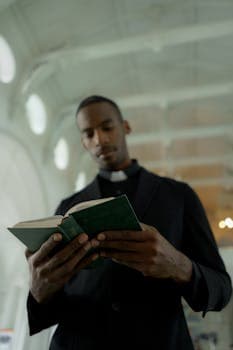 A pastor in a dark suit reads a scripture inside a well-lit church with a serene expression.