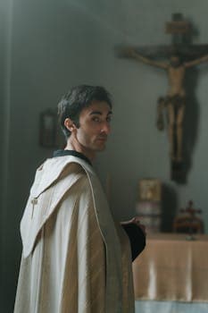 A priest in vestment standing in a softly lit church, looking over his shoulder at a crucifix.