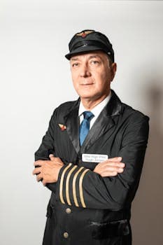 Portrait of a serious airline pilot in uniform with arms crossed against a white background.