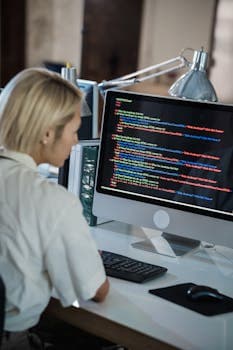 Woman with blonde hair coding on computer in a modern office setting.