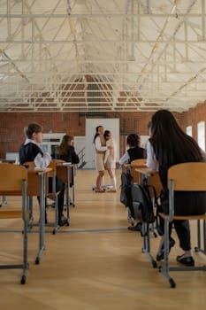 A teacher comforts a student in a bright classroom, observed by other students at their desks.