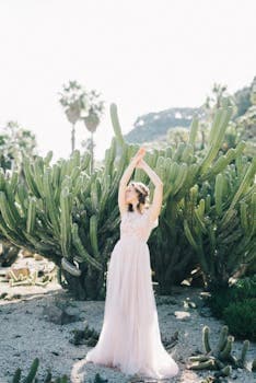 A woman in a flowing white dress poses gracefully among cacti in a sunny garden.