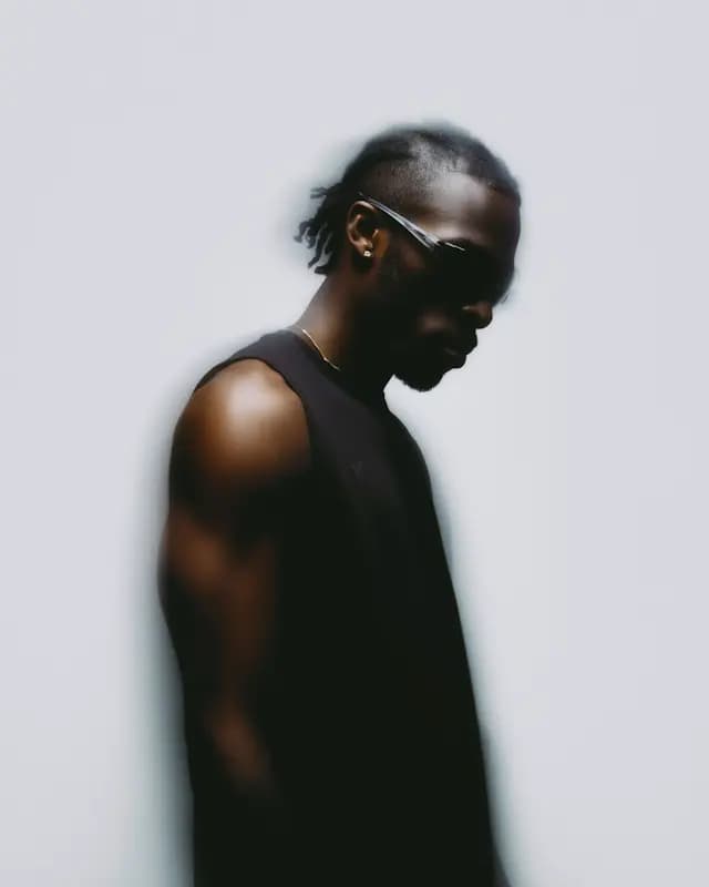 A man wearing a black shirt and sunglasses is standing against a white background. He appears to be looking down, possibly focusing on something or someone. The man's outfit and the contrasting white background create a striking visual effect.