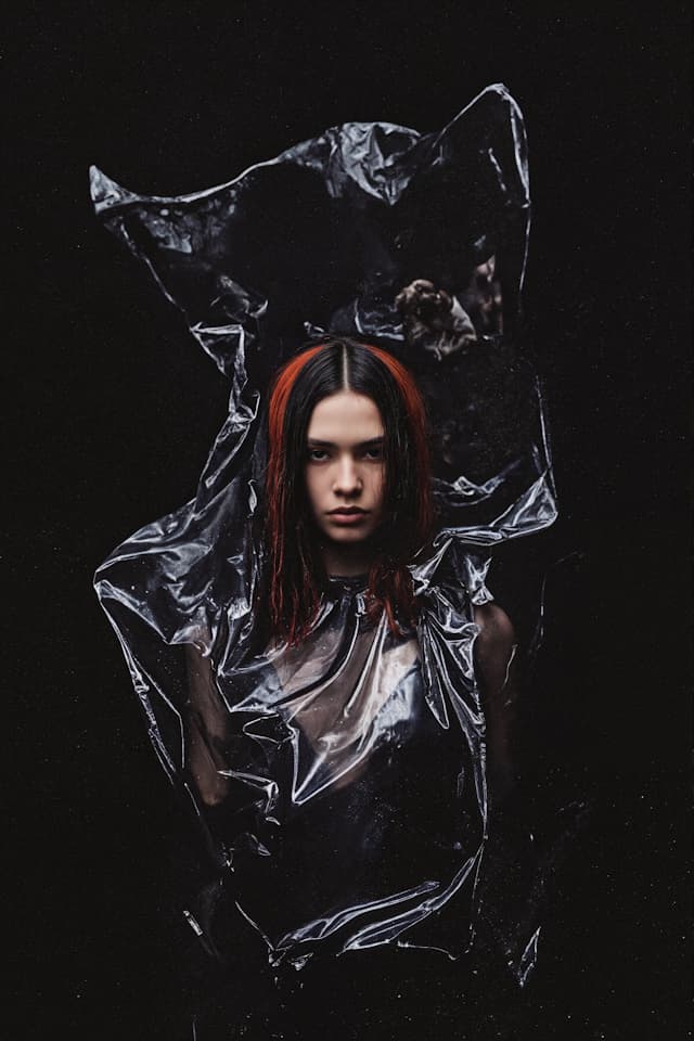 A woman with red hair is standing in front of a black background. She is wearing a black dress and has a red streak in her hair. The woman is positioned in such a way that her head is cut off by the frame, and she appears to be looking at the camera.