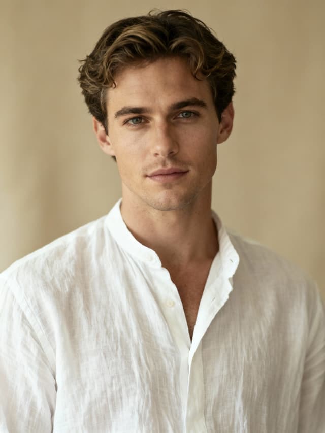 A man with curly hair and a beard is wearing a white shirt and looking at the camera. He has a strong jawline and is posing for the picture. The man's shirt is unbuttoned, revealing his collar.